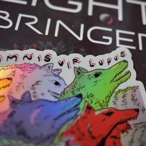 Peut inclure: Un autocollant holographique repr&eacute;sentant une meute de loups stylis&eacute;s de diff&eacute;rentes couleurs : violet, rouge, vert et iris&eacute;. L'autocollant porte l'inscription "OMNIS VIR LUPUS" et le mot "LIGHT BRINGER" en blanc.