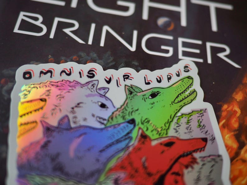 Peut inclure: Un autocollant holographique repr&eacute;sentant une meute de loups stylis&eacute;s de diff&eacute;rentes couleurs : violet, rouge, vert et iris&eacute;. L'autocollant porte l'inscription "OMNIS VIR LUPUS" et le mot "LIGHT BRINGER" en blanc.