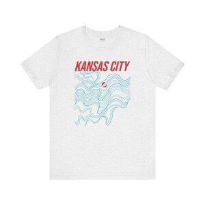 May include: A heather grey t-shirt with a graphic design of blue wavy lines and the text "KANSAS CITY" in red.