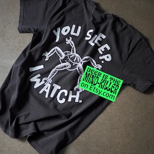 May include: Dark gray t-shirt with a white graphic of a frog-like creature and the words "YOU SLEEP. I WATCH." The shirt also has a green sticker that says "BEER IS THE MIND-KILLER on Etsy.com".