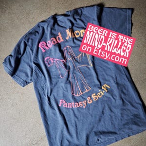 May include: A dark blue t-shirt with the words "Read More" and "Fantasy & Sci-Fi" in pink and orange lettering. The shirt also features a graphic and a sticker that says "Beer is the Mind-Killer on Etsy.com."