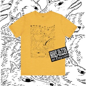 May include: Mustard yellow t-shirt with a black and white wolf illustration. The shirt features the Latin phrase "Omnis Vir Lupus" and a graphic of multiple wolf heads. A black and white sticker with the text "Beer is the Mind Killer" is on the bottom right.