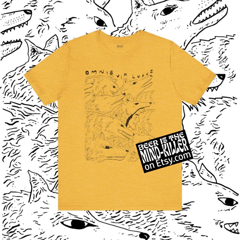 May include: Mustard yellow t-shirt with a black and white wolf illustration. The shirt features the Latin phrase "Omnis Vir Lupus" and a graphic of multiple wolf heads. A black and white sticker with the text "Beer is the Mind Killer" is on the bottom right.