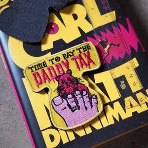 May include: A yellow embroidered patch with a black border featuring a pink rodent graphic and the text "TIME TO PAY THE DADDY TAX" in black and pink. A clear plastic ruler is placed next to the patch, showing a length of approximately 3 inches.