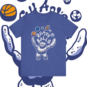 May include: Blue t-shirt with a graphic of a stone-like figure juggling various sports balls. The text "Ghan's Balls" is above the figure. A basketball and other sports-related graphics are in the background.