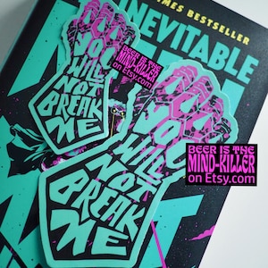May include: Two stickers with a fist graphic and the words "YOU WILL NOT BREAK ME" in teal and pink. The stickers are on a book with the title "THIS INEVITABLE" and the text "NEW YORK TIMES BESTSELLER". A small sticker reads "BEER IS THE MIND-KILLER on Etsy.com".