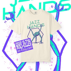 May include: Cream-colored t-shirt with the text "JAZZ HANDS" in teal and a teal and purple graphic of a creature. A purple sticker with the text "BEER IS THE MIND-KILLER on Etsy.com" is on the shirt.