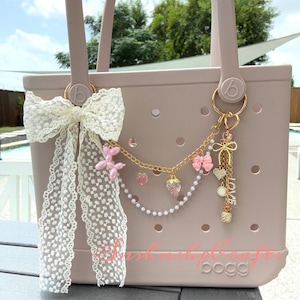 May include: A light pink tote bag with a white lace bow and a gold charm chain. The chain features pink bows, strawberry charms, and a 'LOVE' pendant. The bag has a perforated design.
