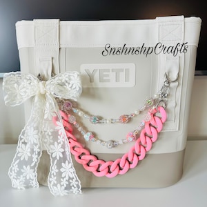 Könnte beinhalten: Eine hellgraue YETI-Tragetasche mit weißen Griffen und Zierleisten. Eine rosa Kette und Perlenaccessoires hängen an der Tasche. Eine weiße Spitzenschleife ist an der Tasche befestigt. Der Text "SnshnshpCrafts" ist auf die Tasche gedruckt.