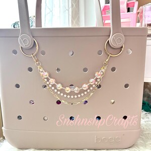 Puede incluir: Un bolso tote de color lila claro con una cadena decorativa. La cadena es de color dorado y presenta cuentas transparentes y rosas, con algunos acentos morados y azules. La bolsa tiene el texto "Snshnshp Crafts" y "bogg" impreso.