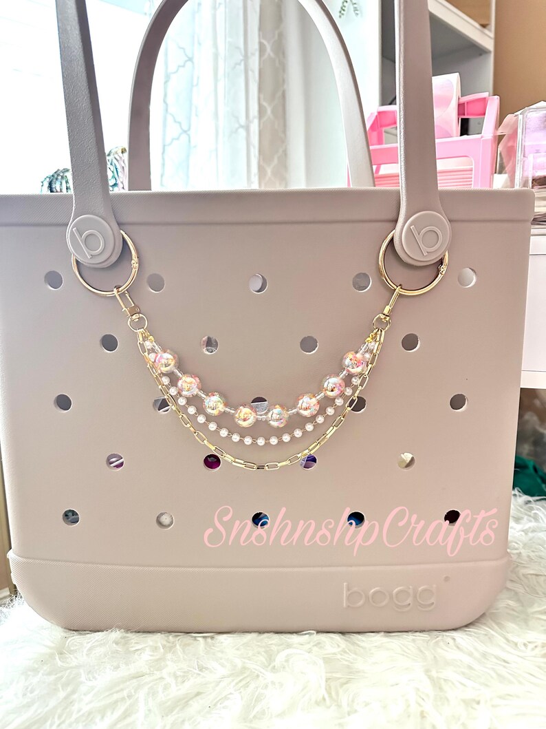 Puede incluir: Un bolso tote de color lila claro con una cadena decorativa. La cadena es de color dorado y presenta cuentas transparentes y rosas, con algunos acentos morados y azules. La bolsa tiene el texto "Snshnshp Crafts" y "bogg" impreso.