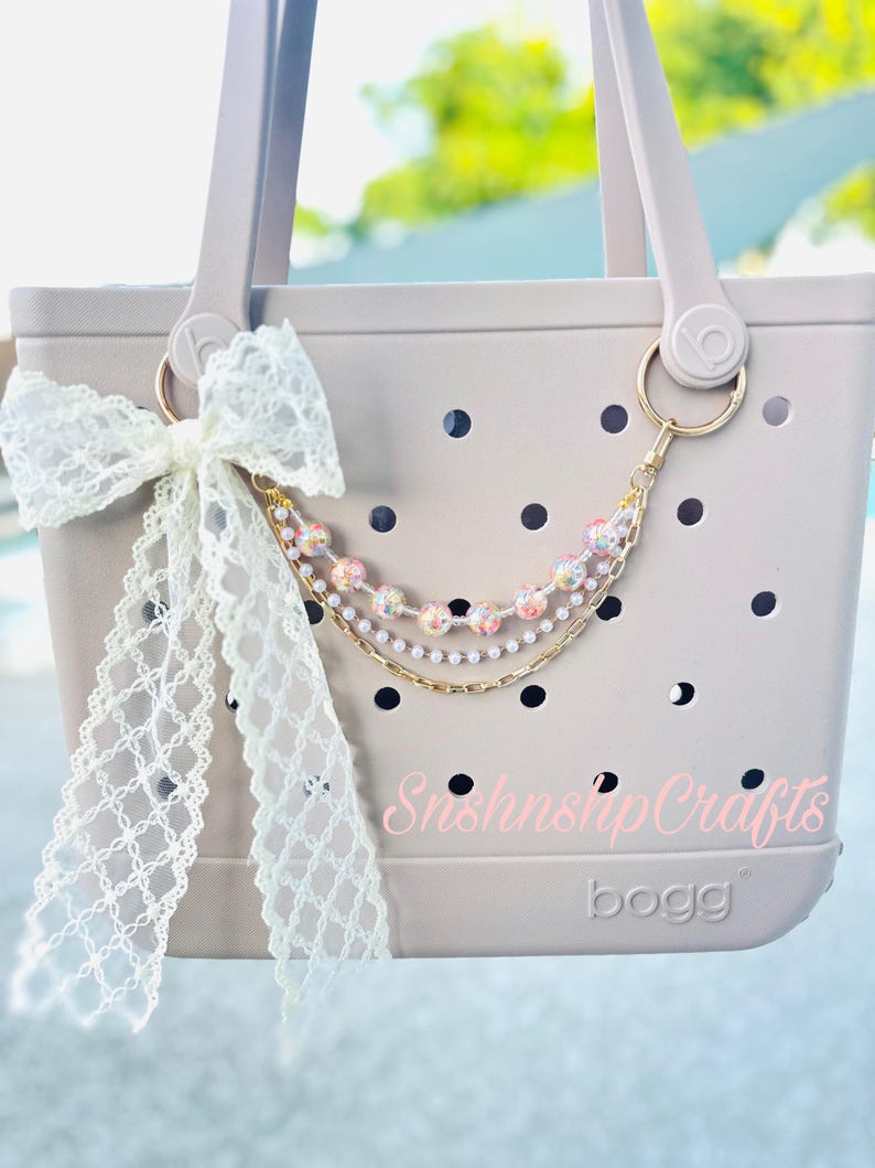 Puede incluir: Un bolso tote beige claro con recortes circulares negros, un lazo de encaje blanco y una cadena dorada decorativa con cuentas rosas y blancas. El bolso tiene un anillo dorado y el texto "SnshnshpCrafts" y "bogg".
