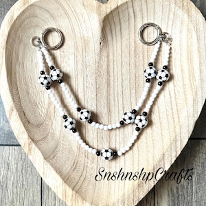 May include: A white beaded lanyard with soccer ball accents and silver-tone clasps. The lanyard features black and white soccer ball beads and is displayed on a wooden heart-shaped tray. The text "SnshnshpCrafts" is visible.