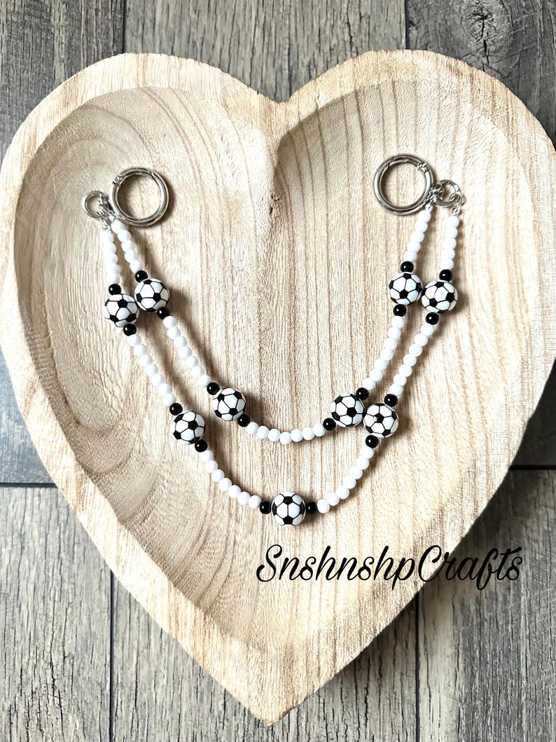 May include: A white beaded lanyard with soccer ball accents and silver-tone clasps. The lanyard features black and white soccer ball beads and is displayed on a wooden heart-shaped tray. The text "SnshnshpCrafts" is visible.