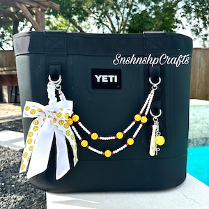 May include: Black YETI cooler bag with a white and yellow bow, and beaded charms. The charms include baseballs, a baseball bat, and a softball. The text "SnshnshpCrafts" is written on the bag.