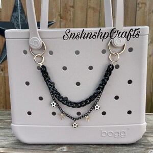 May include: A light gray tote bag with a black chain accessory. The chain features soccer ball charms and gold-colored accents. The bag has a perforated design and the text "SnshnshpCrafts" is visible.