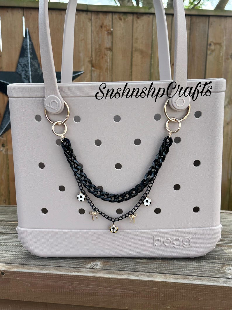 May include: A light gray tote bag with a black chain accessory. The chain features soccer ball charms and gold-colored accents. The bag has a perforated design and the text "SnshnshpCrafts" is visible.