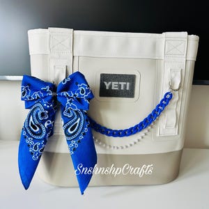 May include: A white and tan cooler bag with a blue paisley bandana bow and a blue chain with pearls. The cooler bag has the brand name "YETI" on the front. The text "SnshnshpCrafts" is printed on the cooler bag.