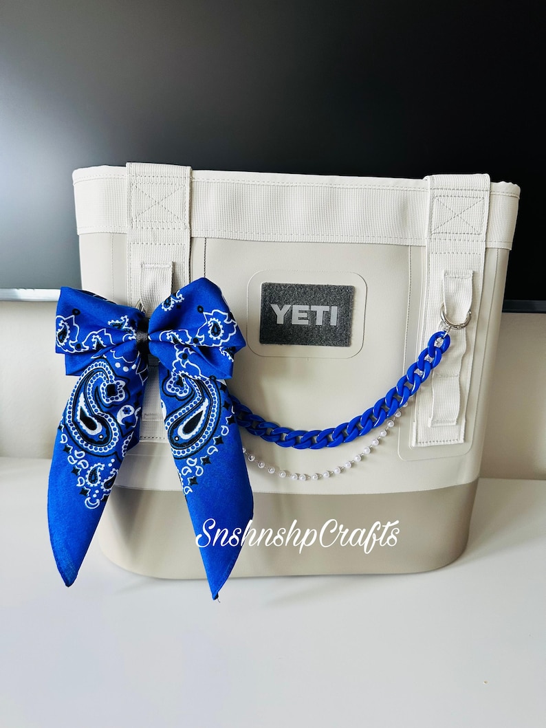 May include: A white and tan cooler bag with a blue paisley bandana bow and a blue chain with pearls. The cooler bag has the brand name "YETI" on the front. The text "SnshnshpCrafts" is printed on the cooler bag.