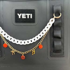 May include: Black cooler bag with a white "YETI" patch. A two-strand chain accessory hangs from the bag. The top chain is white, and the bottom is gold with red basketball charms and the number 17.
