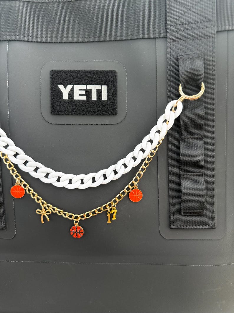 May include: Black cooler bag with a white "YETI" patch. A two-strand chain accessory hangs from the bag. The top chain is white, and the bottom is gold with red basketball charms and the number 17.
