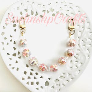 Puede incluir: Una correa de bolso con cuentas con cierres dorados y cuentas transparentes. La correa presenta cuentas redondas transparentes llenas de flores secas en tonos rosa, blanco y amarillo. El texto "SnshnshpCrafts" es visible en rosa.