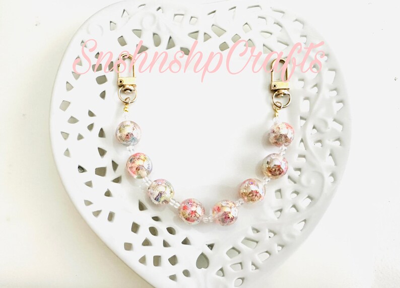Puede incluir: Una correa de bolso con cuentas con cierres dorados y cuentas transparentes. La correa presenta cuentas redondas transparentes llenas de flores secas en tonos rosa, blanco y amarillo. El texto "SnshnshpCrafts" es visible en rosa.