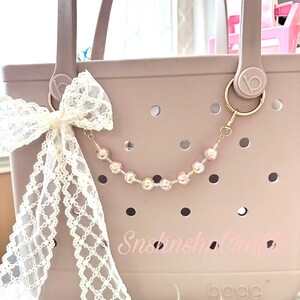 Puede incluir: Un bolso tote malva con un lazo de encaje blanco y un dije de cuentas. El dije presenta cuentas iridiscentes. El bolso tiene un llavero dorado y el texto "Snshnshop Crafts" es visible en el bolso.