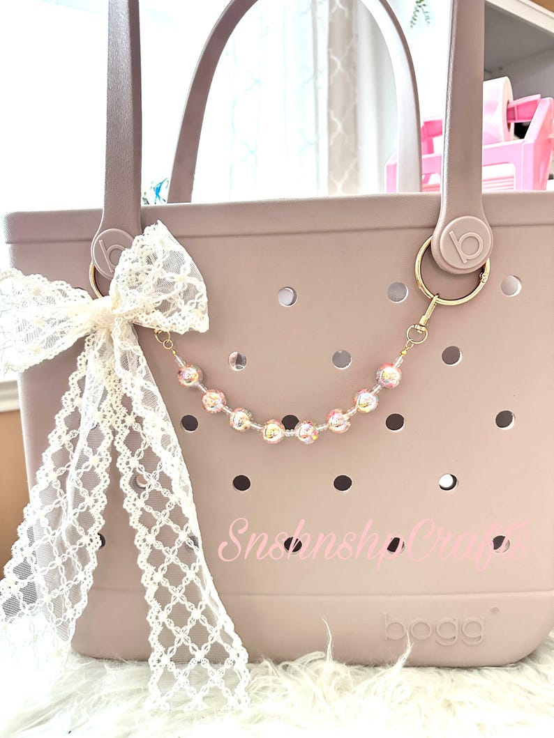 Puede incluir: Un bolso tote malva con un lazo de encaje blanco y un dije de cuentas. El dije presenta cuentas iridiscentes. El bolso tiene un llavero dorado y el texto "Snshnshop Crafts" es visible en el bolso.