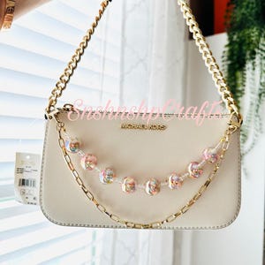 Puede incluir: Un bolso Michael Kors color crema con una correa de cadena dorada y una cadena de cuentas decorativa. La cadena de cuentas presenta cuentas iridiscentes y rosas. El bolso es sostenido por una persona.