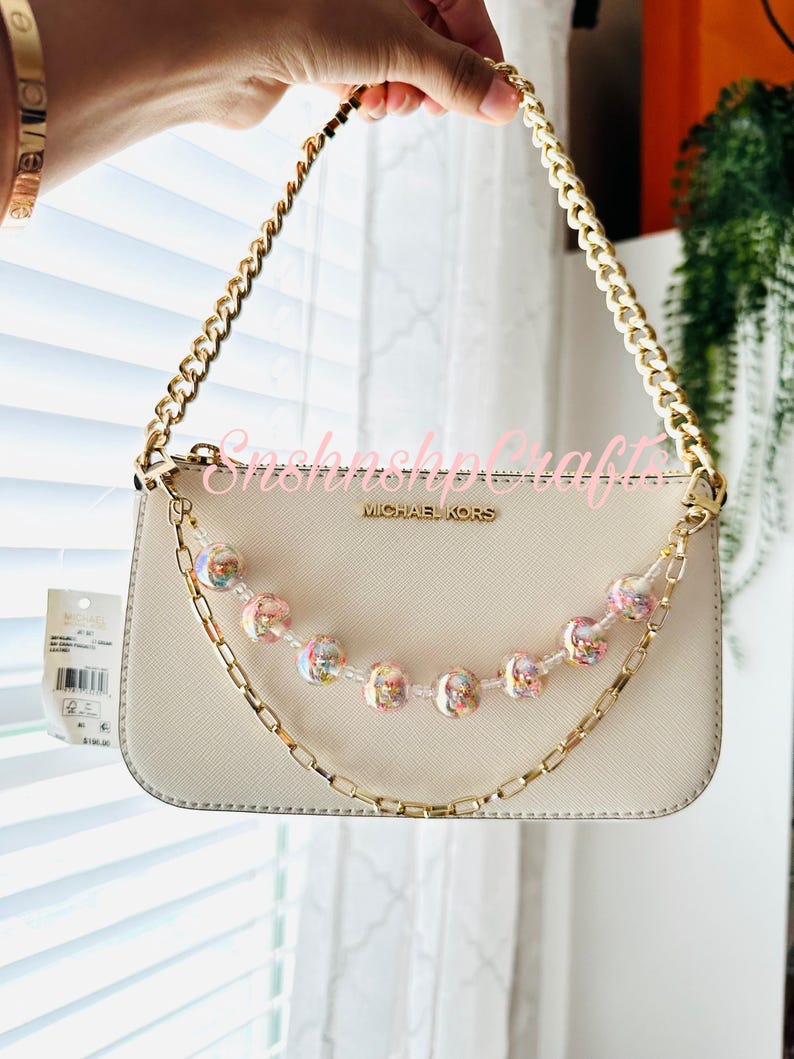 Puede incluir: Un bolso Michael Kors color crema con una correa de cadena dorada y una cadena de cuentas decorativa. La cadena de cuentas presenta cuentas iridiscentes y rosas. El bolso es sostenido por una persona.