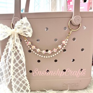 Puede incluir: Un bolso tote color topo con un llavero dorado y un dije de cuentas. Un lazo de encaje blanco est&aacute; sujeto al asa. La bolsa tiene el texto "SnshnshpCrafts" en rosa.