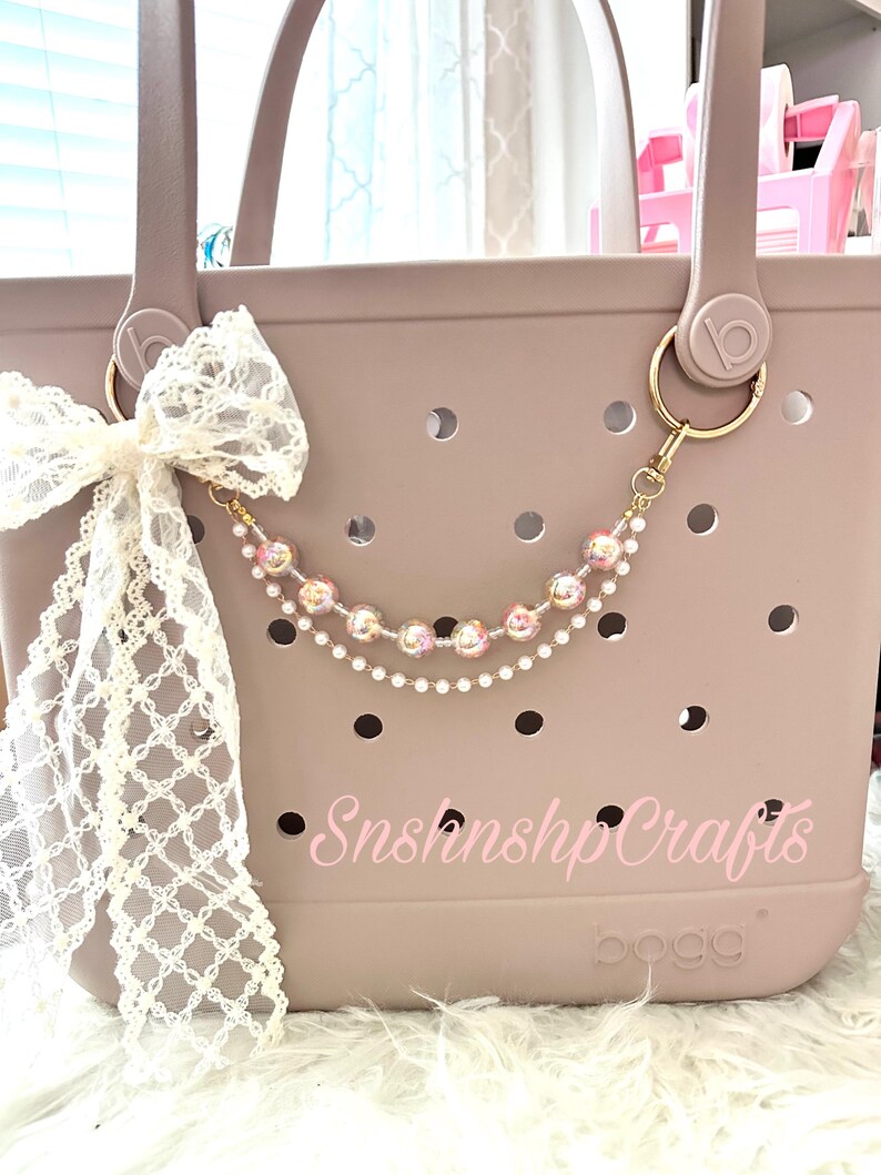 Puede incluir: Un bolso tote color topo con un llavero dorado y un dije de cuentas. Un lazo de encaje blanco est&aacute; sujeto al asa. La bolsa tiene el texto "SnshnshpCrafts" en rosa.