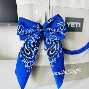 May include: A blue bandana bow with a paisley design is attached to a white cooler. The cooler has a gray "YETI" patch and a blue chain with pearls. The text "Snshnshp Crafts" is visible.