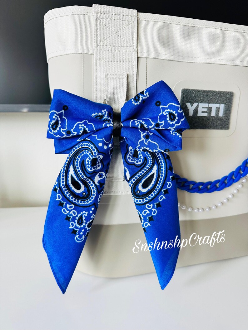 May include: A blue bandana bow with a paisley design is attached to a white cooler. The cooler has a gray "YETI" patch and a blue chain with pearls. The text "Snshnshp Crafts" is visible.