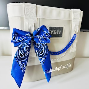 May include: A white and beige cooler bag with a blue paisley bandana bow. The bag has a YETI logo and a decorative chain. The text "Snshnshp Crafts" is visible on the bag.