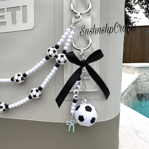 May include: A gray and brown cooler with a soccer-themed keychain. The keychain features a black bow, a small soccer ball, and white beads with soccer ball accents. The text "NAME" is spelled out on the keychain.