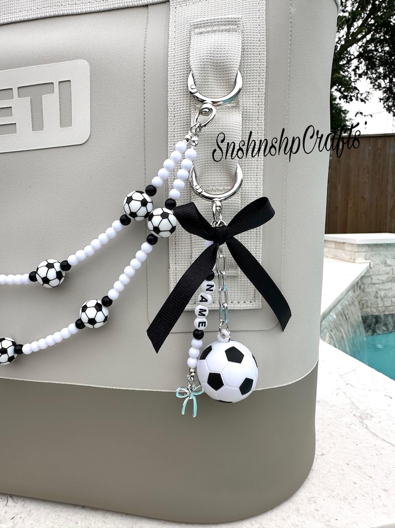 May include: A gray and brown cooler with a soccer-themed keychain. The keychain features a black bow, a small soccer ball, and white beads with soccer ball accents. The text "NAME" is spelled out on the keychain.