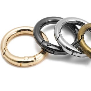 May include: Close-up of four metal spring gate rings in gold, black, silver, and bronze. The rings are circular with a hinged opening and a spring-loaded closure. The rings are designed for attaching items.