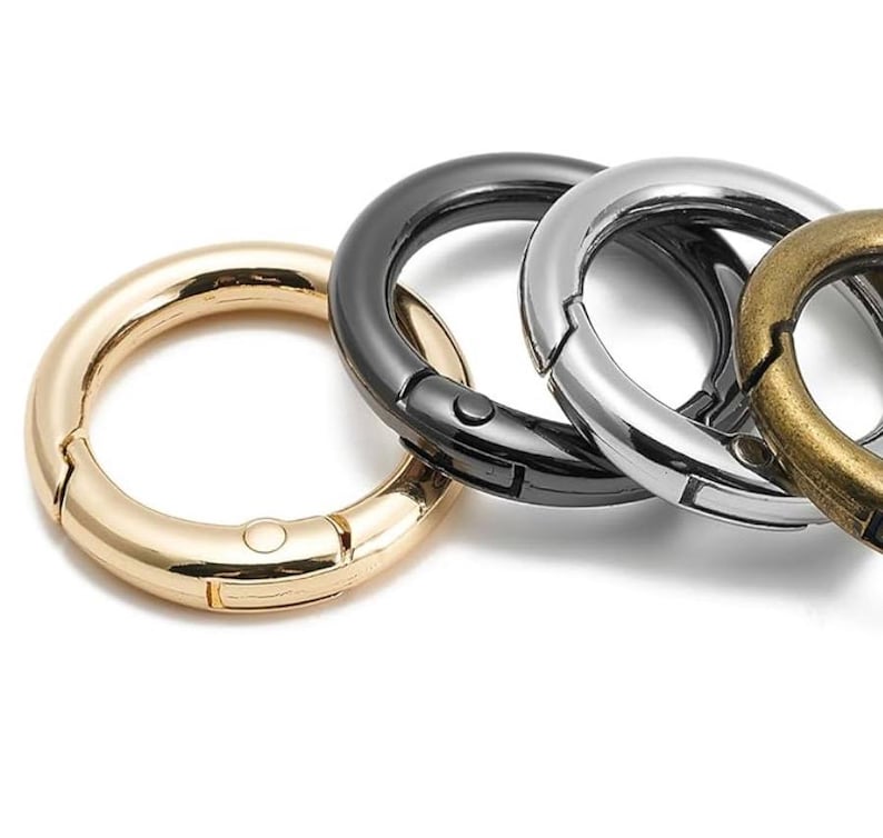 May include: Close-up of four metal spring gate rings in gold, black, silver, and bronze. The rings are circular with a hinged opening and a spring-loaded closure. The rings are designed for attaching items.