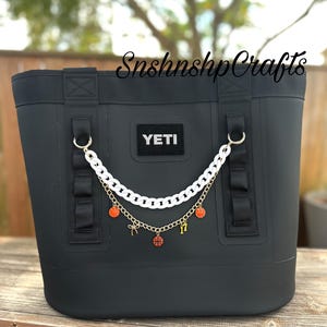 May include: Black YETI tote bag with a white chain and gold chain. The chains have basketball-themed charms. The bag has black handles and a YETI patch. The background is a wooden surface.