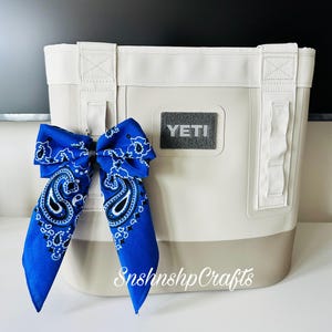 May include: A cream-colored tote bag with a gray base and white straps. A blue paisley bandana is tied to the bag. The word "YETI" is on the front of the bag. The text "SnshnshpCrafts" is on the bottom right.