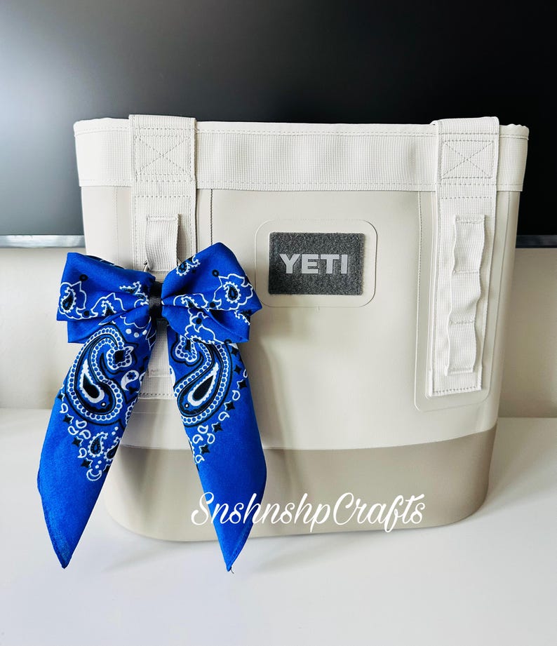 May include: A cream-colored tote bag with a gray base and white straps. A blue paisley bandana is tied to the bag. The word "YETI" is on the front of the bag. The text "SnshnshpCrafts" is on the bottom right.