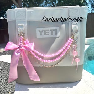 May include: A grey and white YETI cooler with a pink bow and beaded chain. The cooler has the text "SnshnshpCrafts" and "YETI" on the front. Pink lollipop charms hang from the chain.
