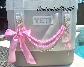 Könnte beinhalten: Grau-weiße YETI-Kühltasche mit rosa Schleife und Perlenkette. Auf der Kühltasche steht "SnshnshpCrafts" und "YETI". Rosa Lutscher-Anhänger hängen an der Kette.