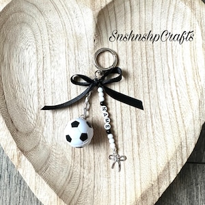 May include: A soccer ball keychain with a black bow and a silver-colored charm. The keychain includes a beaded strand with the name "NAME" spelled out in black and white letter beads. The keychain is on a wooden heart-shaped tray.