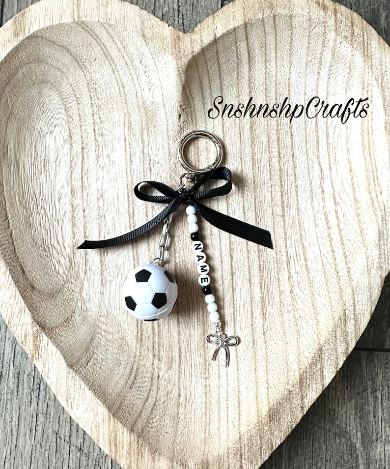 May include: A soccer ball keychain with a black bow and a silver-colored charm. The keychain includes a beaded strand with the name "NAME" spelled out in black and white letter beads. The keychain is on a wooden heart-shaped tray.