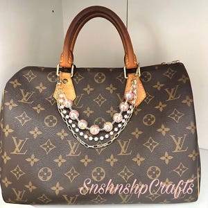 Puede incluir: Bolso marr&oacute;n con un patr&oacute;n de logotipo repetido, asas de cuero marr&oacute;n y herrajes dorados. Una cadena decorativa con cuentas rosas y blancas cuelga de las asas. El texto "Snshnshp Crafts" est&aacute; escrito en rosa.