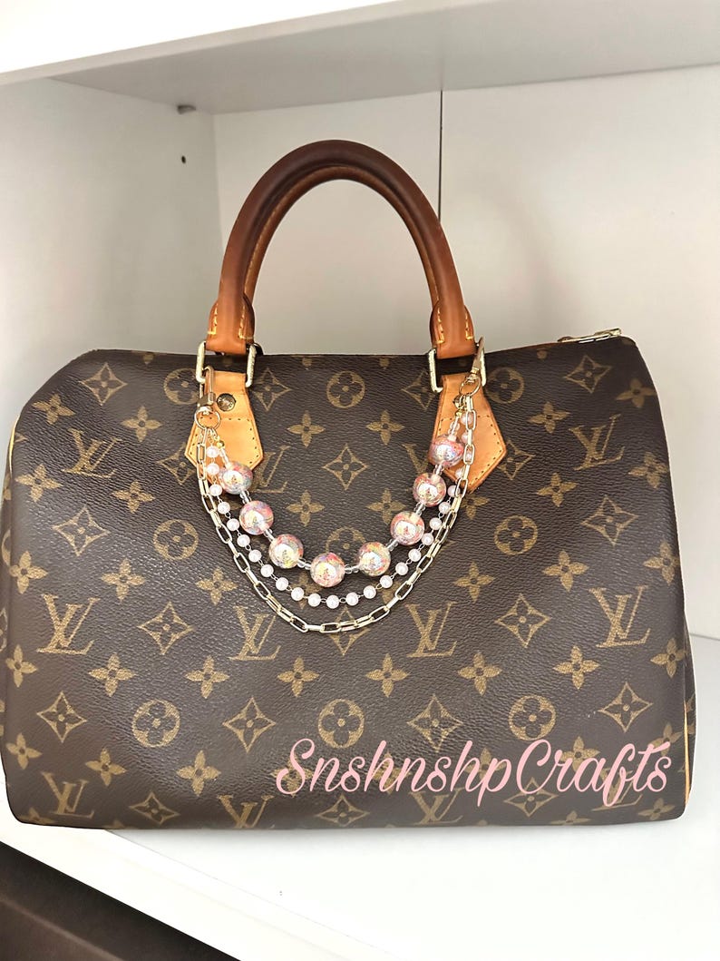 Puede incluir: Bolso marr&oacute;n con un patr&oacute;n de logotipo repetido, asas de cuero marr&oacute;n y herrajes dorados. Una cadena decorativa con cuentas rosas y blancas cuelga de las asas. El texto "Snshnshp Crafts" est&aacute; escrito en rosa.