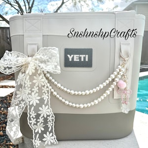 May include: A gray and white YETI cooler bag with a white lace bow and a pearl and charm accessory. The bag has the text "SnshnshpCrafts" on the side.